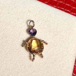 Peut inclure: Pendentif doré en forme de personne. La tête est une pierre en forme de cœur violet, et le corps est une grosse pierre ronde jaune-orangé. De petits cercles dorés forment les bras et les jambes.