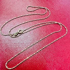 May include: A delicate gold-colored necklace with a small clasp, resting on a textured red surface. The necklace features a series of small, elongated beads strung together to form a chain. The clasp is visible, indicating the necklace's ability to be opened and closed.