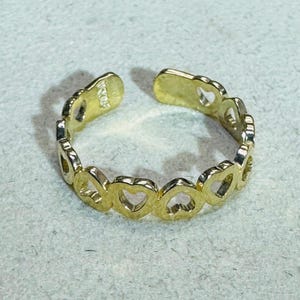Puede incluir: Un anillo abierto de tono dorado que presenta una banda continua de recortes interconectados en forma de corazón. La superficie metálica tiene un acabado pulido y el anillo es ajustable con una pequeña abertura en la parte superior. Tiene un sello.