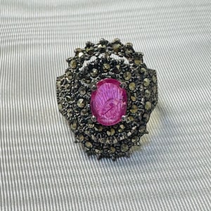 May include: A silver-toned ring with a large, oval, pink gemstone at the center. The ring is surrounded by small, round, dark-colored stones, creating a detailed, textured design. The ring's setting has an ornate, vintage style.