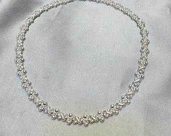 May include: A silver-toned metal necklace featuring a repeating pattern of small, rounded four-petal flower shapes alternating with polished X-shaped links. The necklace is arranged in a circular formation and includes a secure metal clasp closure.