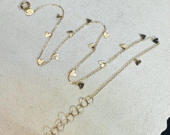 May include: A delicate gold-colored necklace with small heart charms evenly spaced along the chain. The necklace has a lobster clasp closure and an extender chain. The hearts are a warm gold color, and the chain is fine and elegant.