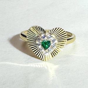 May include: A gold ring featuring a heart-shaped design. The ring has a central emerald-colored stone in a heart shape, surrounded by smaller white stones. The heart is set within a sunburst design.