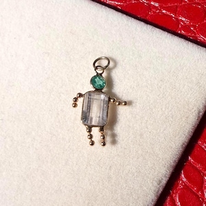 Peut inclure: Pendentif doré en forme de personne. La tête est une pierre précieuse verte, le corps est une pierre précieuse claire rectangulaire et les bras et les jambes sont de petites perles dorées. Le pendentif est sur une surface blanche.