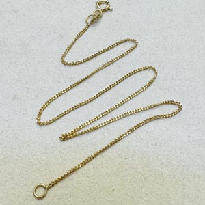 Op de afbeelding: Een fijne gouden ketting bestaande uit een reeks kleine, in elkaar grijpende schakels. De ketting is aan het ene uiteinde afgewerkt met een ronde veerringsluiting en aan het andere uiteinde met een kleine, ronde ring voor een sluiting.