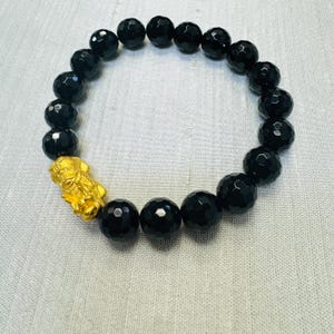 May include: Black onyx beaded bracelet with a gold charm. The beads are faceted and the charm is detailed with intricate designs.