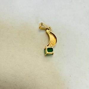 K&ouml;nnte beinhalten: Goldener Anh&auml;nger mit geschwungenem Design und einem quadratischen, smaragdgr&uuml;nen Edelstein am unteren Ende. Der Anh&auml;nger hat oben eine kleine Schlaufe f&uuml;r eine Kette. Der Anh&auml;nger ist vor neutralem Hintergrund.