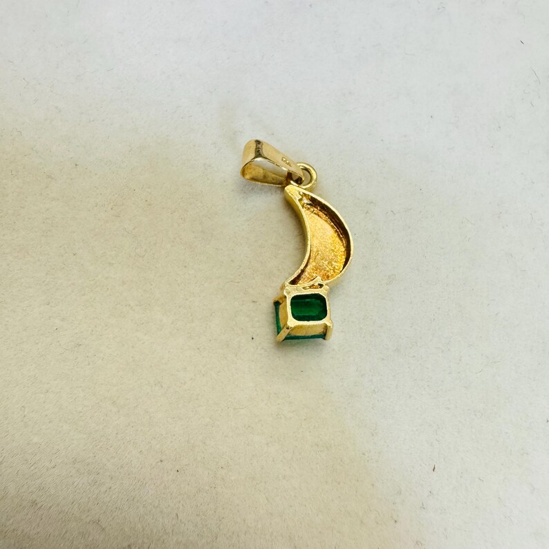 K&ouml;nnte beinhalten: Goldener Anh&auml;nger mit geschwungenem Design und einem quadratischen, smaragdgr&uuml;nen Edelstein am unteren Ende. Der Anh&auml;nger hat oben eine kleine Schlaufe f&uuml;r eine Kette. Der Anh&auml;nger ist vor neutralem Hintergrund.