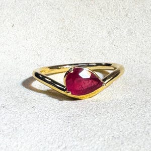Puede incluir: Un anillo de oro con una gema roja oscura en forma de pera. La banda del anillo es de oro pulido, simple y curvada para abrazar la piedra. La piedra roja vibrante es el punto focal, capturando la luz y mostrando su color.