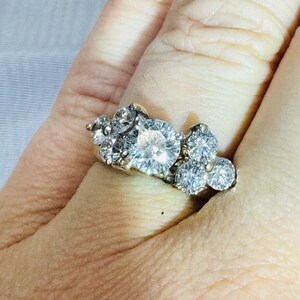 May include: A cluster ring featuring a large, round, clear gemstone surrounded by smaller, clear gemstones. The ring is set in a gold band and is worn on a finger.