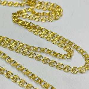 May include: Close-up of multiple gold-colored chain necklaces. The chains have a delicate, intricate design with a repeating pattern of linked loops. The chains are displayed against a light background.