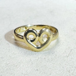 Op de afbeelding: Een goudkleurige ring met een hartvormig ontwerp. Het hart wordt gevormd door twee in elkaar grijpende lussen, waardoor een open midden ontstaat. De band van de ring is glad en eenvoudig, als aanvulling op de gedetailleerde hartvorm.