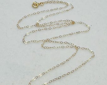May include: Delicate gold-colored chain necklace with a lobster clasp closure. The chain is made of small, interconnected oval links. The necklace is displayed on a light-colored surface, highlighting its subtle shine. A simple and elegant jewelry piece.