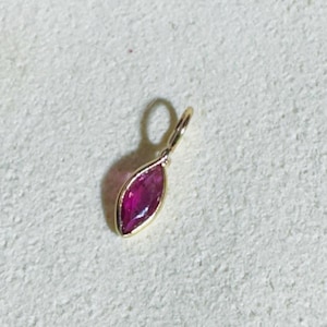 May include: A gold pendant with a marquise-cut, deep red gemstone. The gemstone is set in a gold bezel, and the pendant has a small loop at the top for a chain. The pendant is displayed on a light-colored surface.