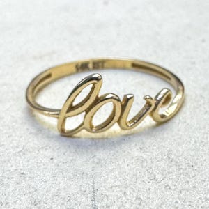 May include: A gold ring featuring the word love in a cursive script font. The band is thin and circular, with the text love integrated into the top section. The interior of the band is clearly stamped with the text 14K JST, indicating the metal purity.