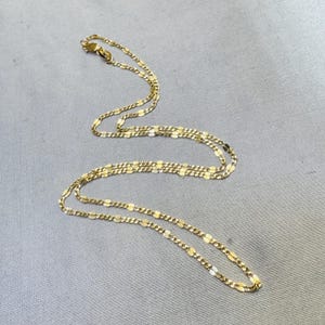 Op de afbeelding: Een goudkleurige ketting bestaande uit een reeks langwerpige, afgeplatte ovale schakels die in een herhalend patroon zijn verbonden. De ketting is voorzien van een kleine, ronde karabijnsluiting aan één uiteinde met een gepolijste afwerking.