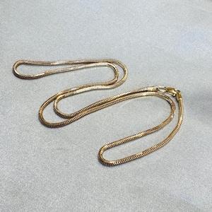 Puede incluir: Un collar de cadena tipo serpiente en tono dorado con una estructura de eslabones metálicos lisos, flexibles y estrechamente tejidos. La cadena está dispuesta en un bucle curvo suelto e incluye un pequeño cierre de mosquetón funcional.