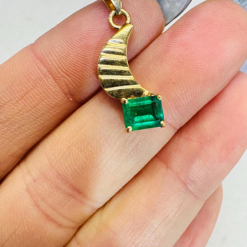 K&ouml;nnte beinhalten: Goldener Anh&auml;nger mit einem halbmondf&ouml;rmigen Design mit horizontalen Linien und einem rechteckigen Smaragd-Edelstein. Der Anh&auml;nger h&auml;ngt an einer kleinen Goldschlaufe.