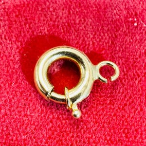 May include: A gold-colored spring ring clasp, a jewelry component, is shown against a red background. The clasp is circular with a small lever and a ball-shaped detail. The metal has a shiny finish.