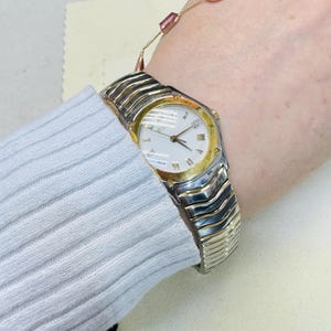 Pode incluir: Um rel&oacute;gio de pulso de duas cores com uma pulseira prateada e uma moldura dourada. O mostrador do rel&oacute;gio &eacute; branco com ponteiros pretos e algarismos romanos. O rel&oacute;gio est&aacute; em um pulso, com uma manga de su&eacute;ter branca vis&iacute;vel.