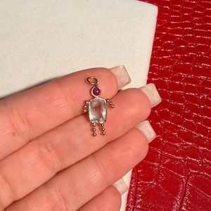 Peut inclure: Un petit charme doré en forme de personne. Le charme présente un corps rectangulaire en pierre transparente, une tête ronde en pierre rouge et de petits accents de boules dorées pour les bras et les jambes. Le charme est tenu par une main.