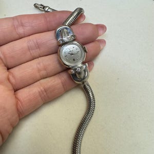 K&ouml;nnte beinhalten: Eine silberne Armbanduhr mit wei&szlig;em Zifferblatt und Diamantbesatz am Band. Die Uhr hat ein Schlangenkettenband.