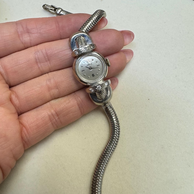 K&ouml;nnte beinhalten: Eine silberne Armbanduhr mit wei&szlig;em Zifferblatt und Diamantbesatz am Band. Die Uhr hat ein Schlangenkettenband.