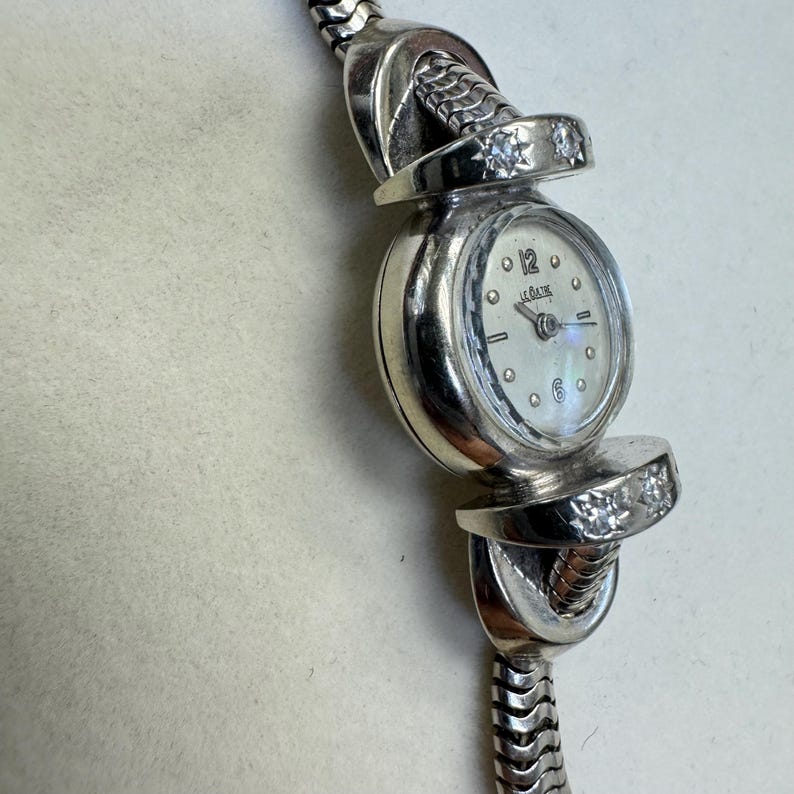 K&ouml;nnte beinhalten: Eine silberne Armbanduhr mit wei&szlig;em Zifferblatt und Diamant-Akzenten am Armband. Das Zifferblatt der Uhr hat schwarze r&ouml;mische Ziffern als Stundenmarkierungen und den Text "Le Coultre" aufgedruckt.
