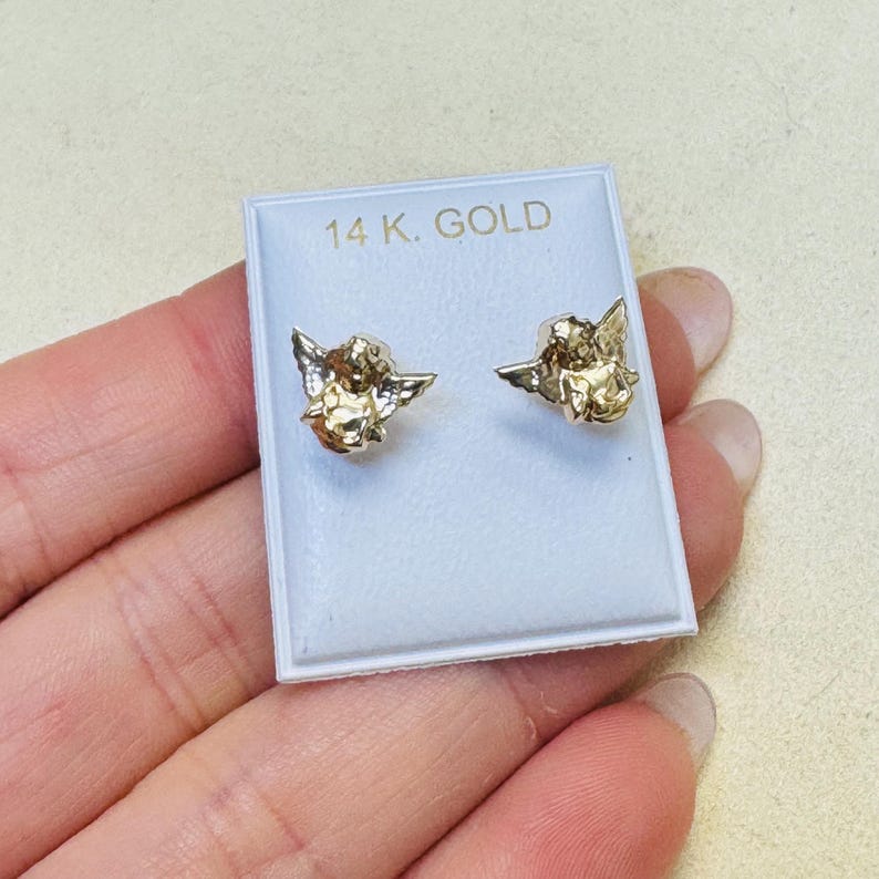 Op de afbeelding: Een paar goudkleurige engel-oorstekers op een witte displaykaart. De kaart heeft de tekst "14 K. GOLD" in zwarte letters. De oorbellen zijn klein, gedetailleerd en hebben gevleugelde cherubijnontwerpen.