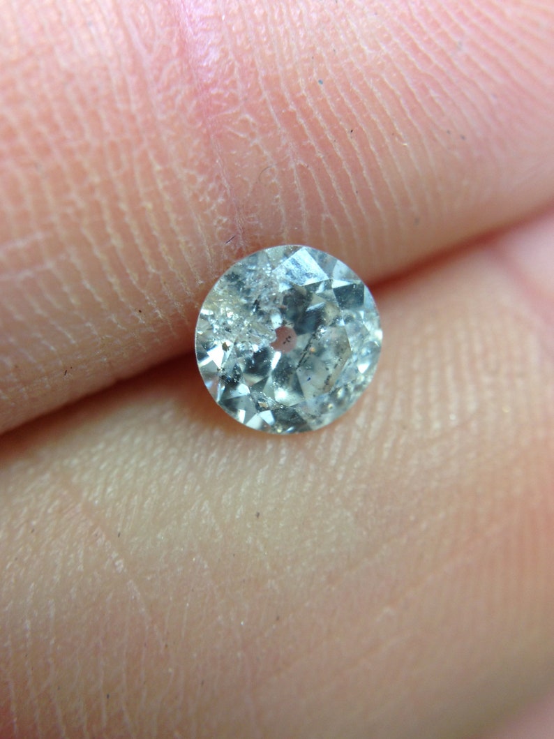 May include: A clear, round, faceted gemstone with a slight blue hue. The gemstone is set in a silver bezel.