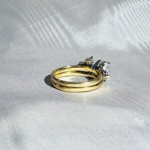 May include: A gold wedding ring set with a diamond engagement ring. The rings are on a white satin surface. The engagement ring has a large, clear diamond and smaller accent stones.