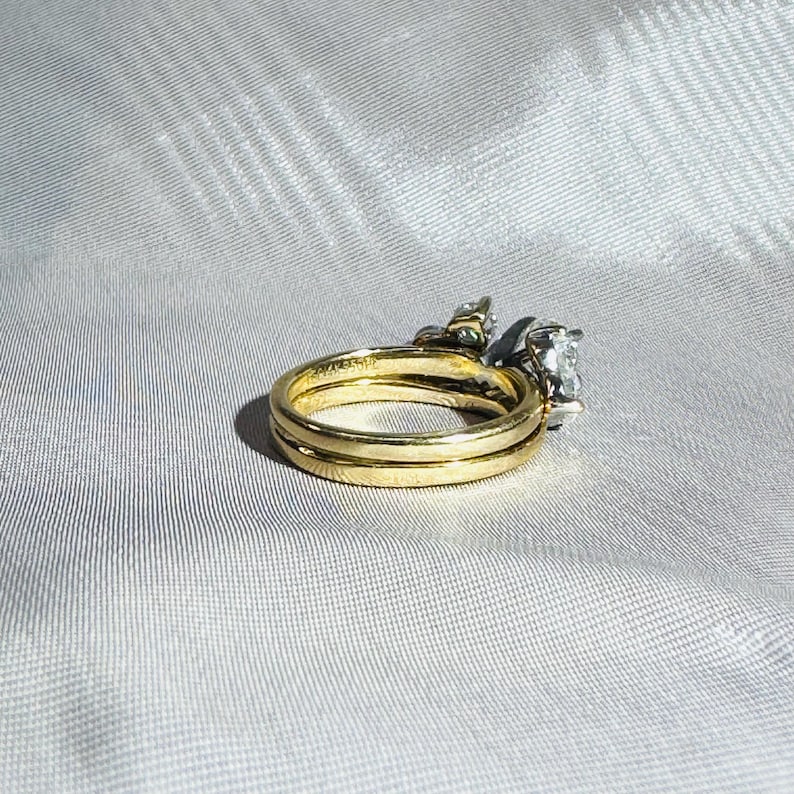 May include: A gold wedding ring set with a diamond engagement ring. The rings are on a white satin surface. The engagement ring has a large, clear diamond and smaller accent stones.