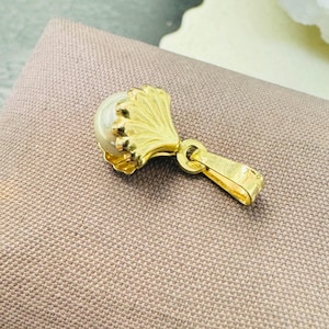 May include: A gold pendant featuring a pearl set in a shell-shaped design. The pendant has a loop for attaching to a chain.