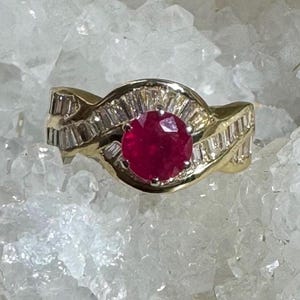 Op de afbeelding: Een gouden ring met een grote, ronde, rode edelsteen in het midden, omringd door kleinere, rechthoekige, heldere edelstenen. De band heeft een wervelend ontwerp. De ring is geplaatst tegen een witte, kristallijne achtergrond.