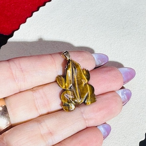 May include: A detailed frog pendant carved from tiger's eye gemstone. The pendant features a gold-colored bail for attaching to a chain. The frog is a rich, golden brown color with natural patterns.