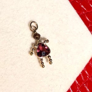 Peut inclure: Pendentif en forme de personnage, avec une grosse pierre rose foncé triangulaire pour le corps. La tête est une petite pierre brune rectangulaire. Des perles dorées forment les bras et les jambes. Le pendentif est sur une surface blanche.