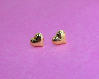 May include: Two small, gold-colored heart-shaped stud earrings. The earrings are positioned on a pink background. The hearts have a smooth, reflective surface, catching the light. The earrings are a classic jewelry item.