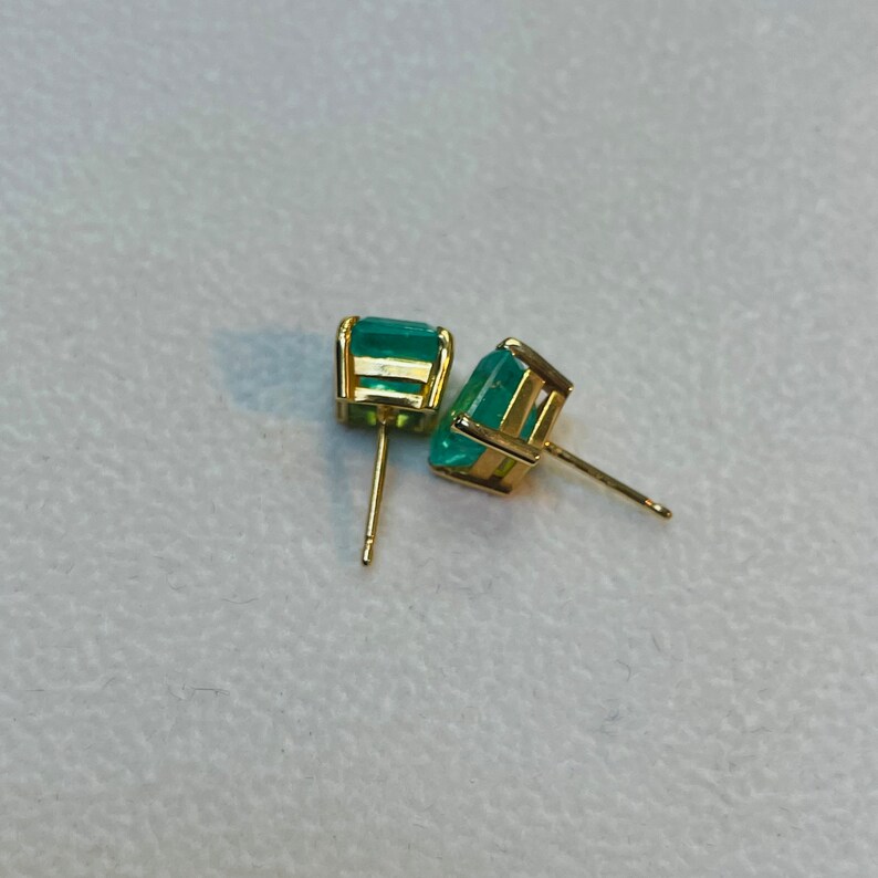 May include: A pair of gold earrings with emerald green gemstones. The earrings are square shaped and have a simple design.