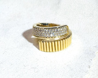 May include: A gold-toned ring with a wrap-around design. The top band is encrusted with small, sparkling stones, while the lower band features vertical, ridged detailing. The ring is set against a white background.