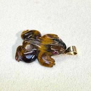 May include: A carved tiger's eye frog pendant with a gold bail and a small red gemstone eye. The frog is brown and gold with detailed carving. The pendant is a unique piece of jewelry.