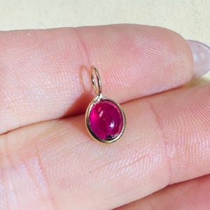 Puede incluir: Un pequeño colgante ovalado con una vibrante piedra preciosa de color rojo rubí engastada en un marco de metal dorado. El colgante tiene un lazo en la parte superior para sujetarlo a una cadena. La piedra es lisa y pulida.