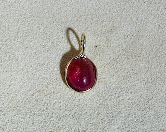 May include: A gold pendant with a large, oval, deep red gemstone. The gemstone is set in a gold bezel and has a gold loop for a chain. The pendant is displayed on a textured, light beige surface.