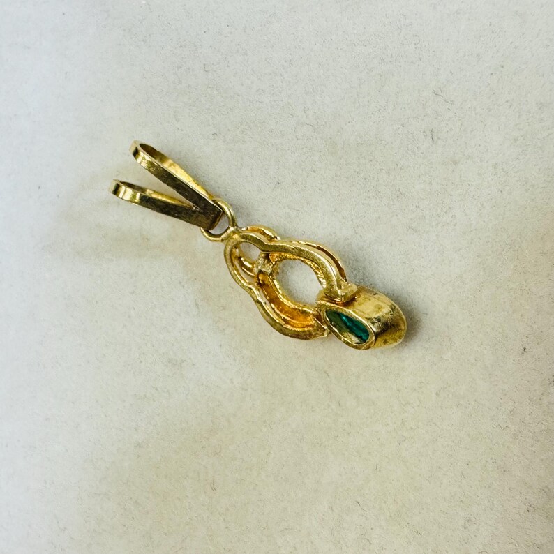 May include: A gold-colored pendant with a clasp and a small, oval-shaped emerald. The pendant has a decorative, looped design and is set against a light background.