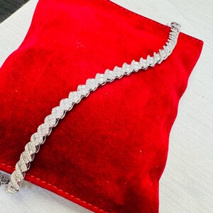 May include: A silver bracelet with a repeating, wave-like pattern of clear gemstones. The bracelet is displayed on a red velvet surface.