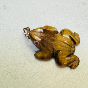 May include: A carved frog pendant made of tiger's eye gemstone. The pendant is brown with gold streaks and a small red gemstone eye. It has a silver-colored bail for attaching to a chain.