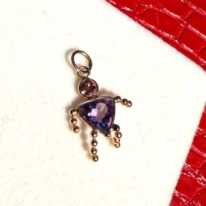 Peut inclure: Un pendentif doré en forme de personne. La tête est une pierre précieuse rose, le corps est une pierre précieuse violette triangulaire et les bras et les jambes sont de petites perles dorées. Le pendentif a une boucle en haut pour une chaîne.