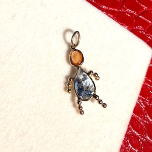 Peut inclure: Un pendentif doré en forme de personne. La tête est une pierre ovale orange, le corps est une pierre bleue en forme de poire, et les bras et les jambes sont de petites perles dorées. Le pendentif est sur une surface blanche avec un fond rouge.