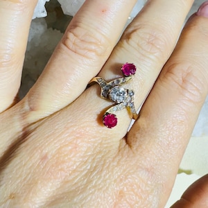 Peut inclure: Une bague en or avec un grand diamant rond et deux accents rubis. La bague a un design unique et tourbillonnant.