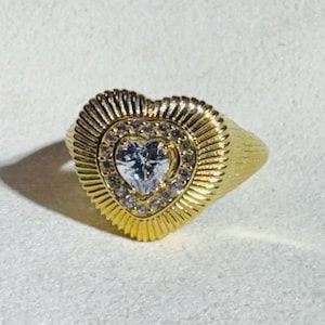 May include: A gold-colored ring with a heart-shaped design. The ring features a central heart-shaped stone surrounded by smaller stones. The band has a textured, ridged pattern.