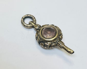 May include: A gold-toned key with an ornate design and a pink glass stone in the center. The key has a loop at the top for attaching to a chain.